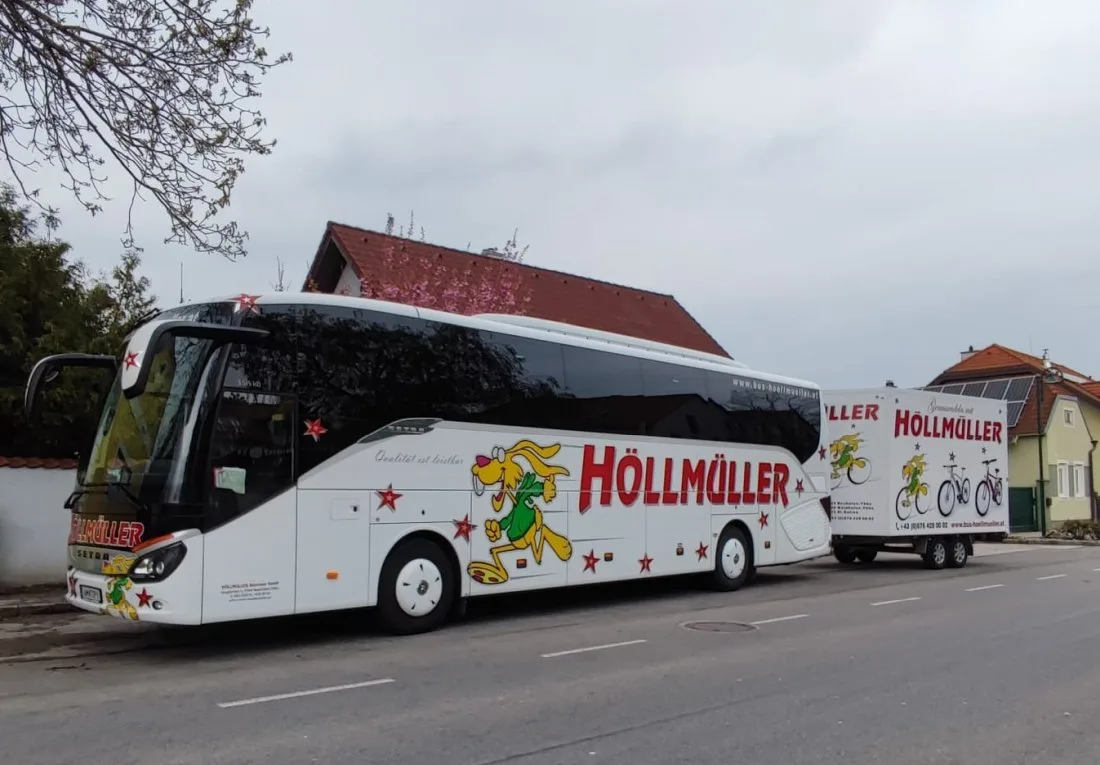 Weißer Höllmüller-Reisebus mit gelbem Hasen und rotem Schriftzug, dahinter der Radanhänger, an einer Ortsstraße unter blühenden Bäumen
