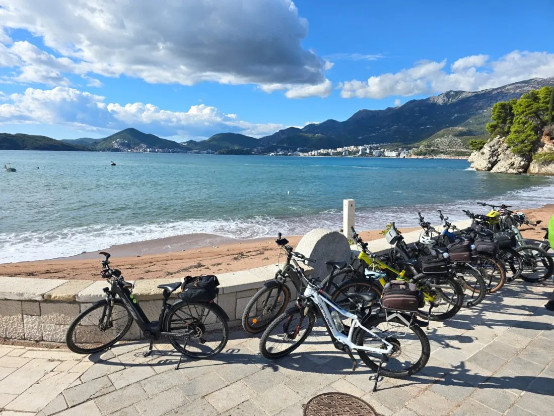 Eine Reihe E-Bikes an einer Strandpromenade, dahinter Meer und Berge