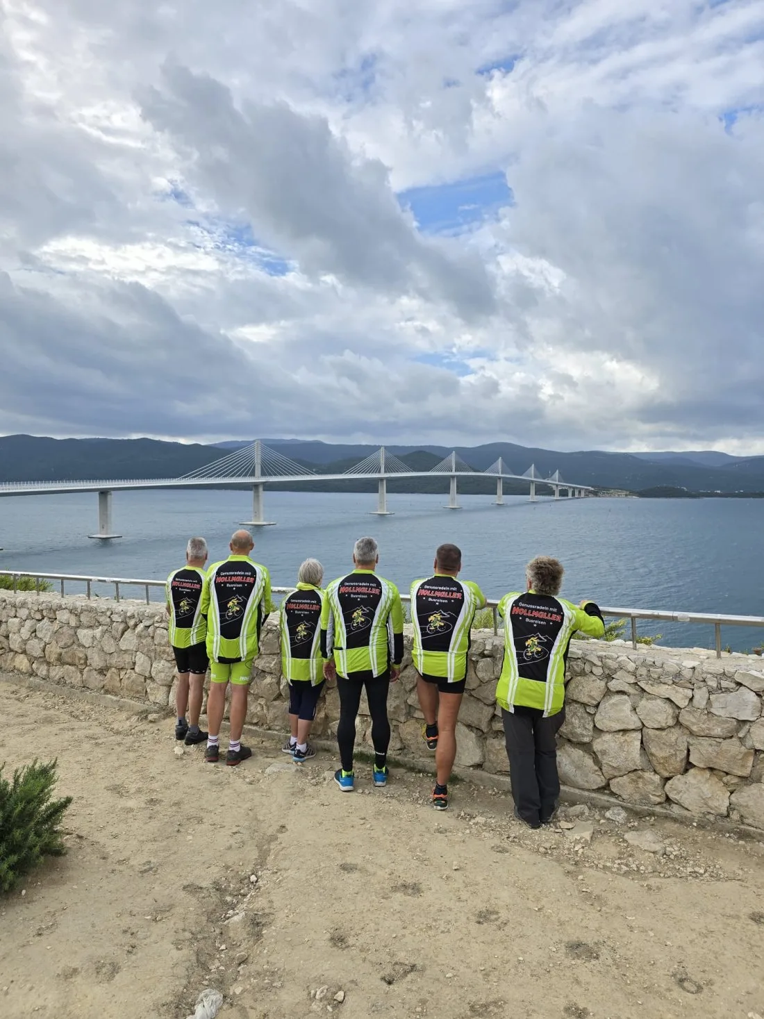 Sechs Radfahrer in grün-schwarzen Höllmüller-Trikots stehen an einer Steinmauer und blicken auf eine lange Schrägseilbrücke über dem Meer
