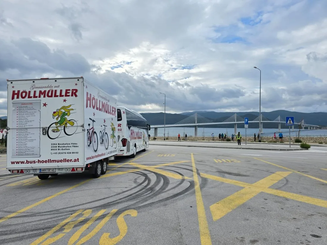 Der Höllmüller-Radanhänger von hinten mit Bus auf einem Parkplatz, im Hintergrund eine Brücke über das Meer