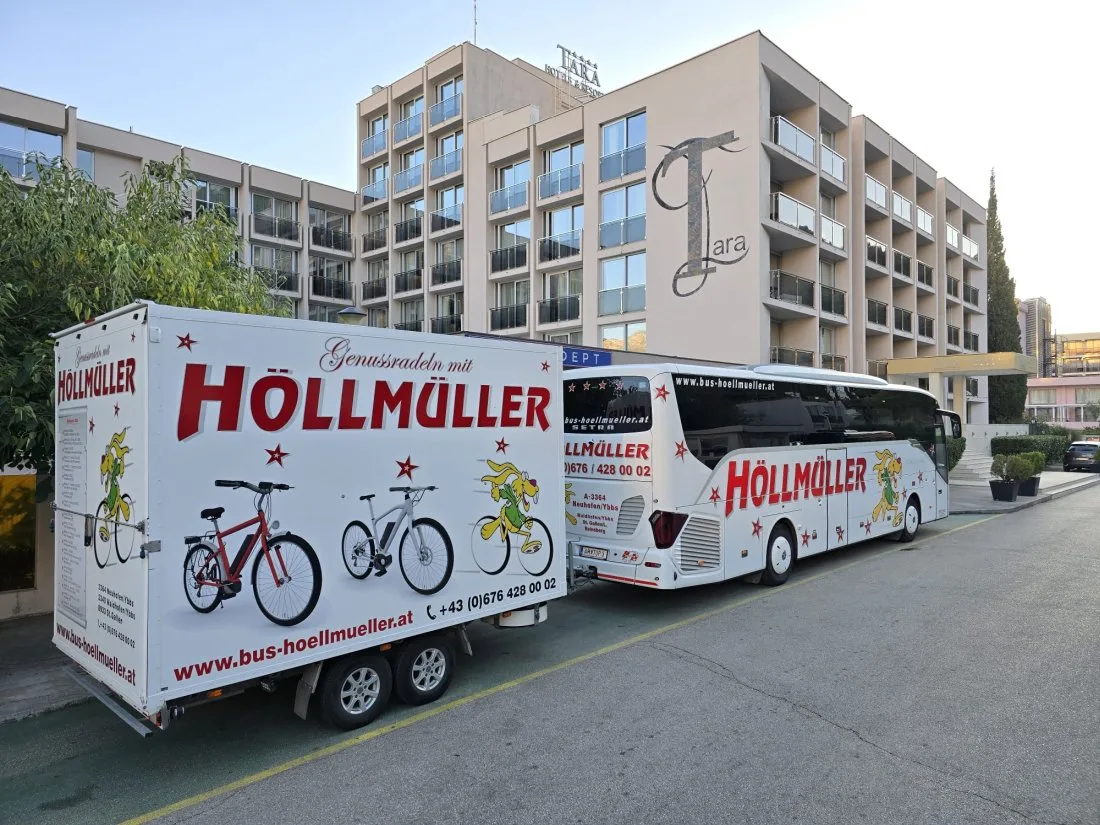 Radanhänger mit der Aufschrift „Genussradeln mit Höllmüller“ und ein Setra-Reisebus vor einem Hotel